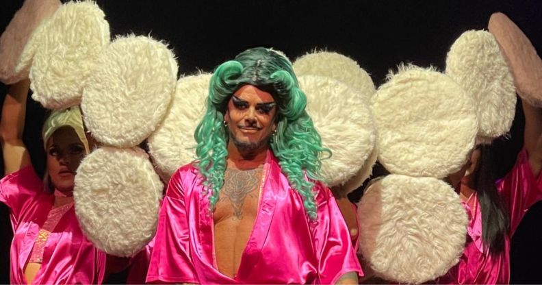 A drag performer in a pink robe and green wig surrounded by large powder puffs.