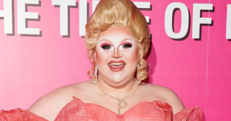 Pixie Polite in a pink dress and blonde wig posing and smiling against a pink and white background.