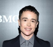 Elliot Page stands against a grey backdrop. He's wearing a grey shirt and tie with a black blazer. Smiling into the camera.