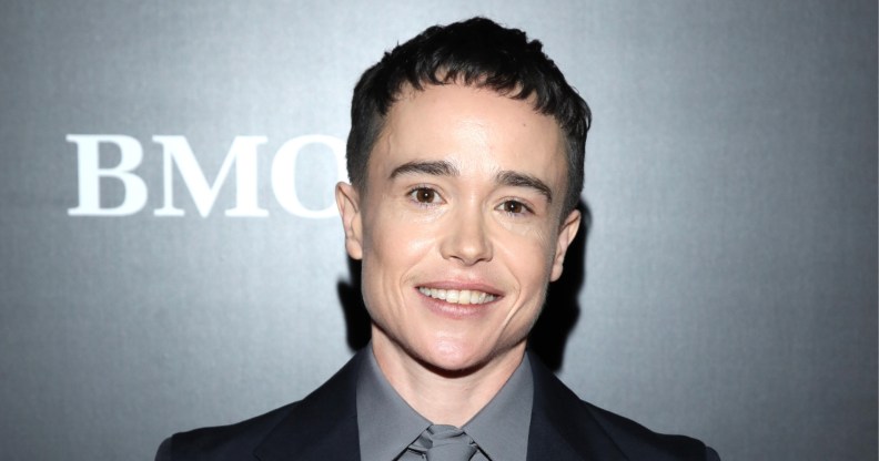 Elliot Page stands against a grey backdrop. He's wearing a grey shirt and tie with a black blazer. Smiling into the camera.
