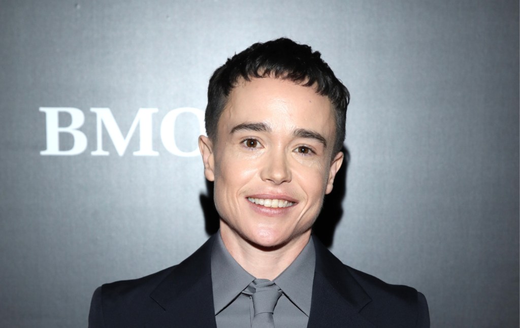 Elliot Page stands against a grey backdrop. He's wearing a grey shirt and tie with a black blazer. Smiling into the camera.
