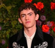 Joe Locke poses in a black and silver jacket and white shirt while stood against a bush and flowers