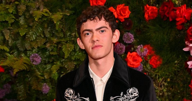 Joe Locke poses in a black and silver jacket and white shirt while stood against a bush and flowers