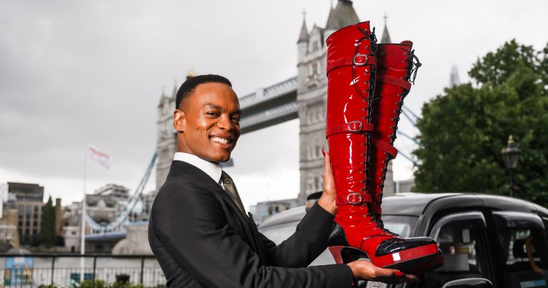 Johannes Radebe in front of London bridge and a black taxi holding up Kinky Boots' red lace hup knee-high boots