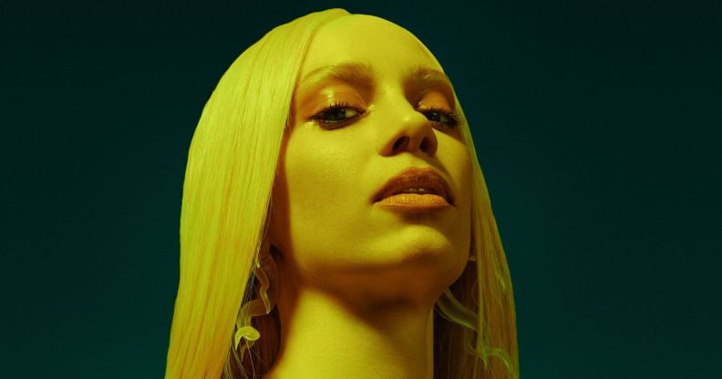 Pop singer Agnes poses in a blonde wig. She is showered in a yellow light, against a blue-black background.