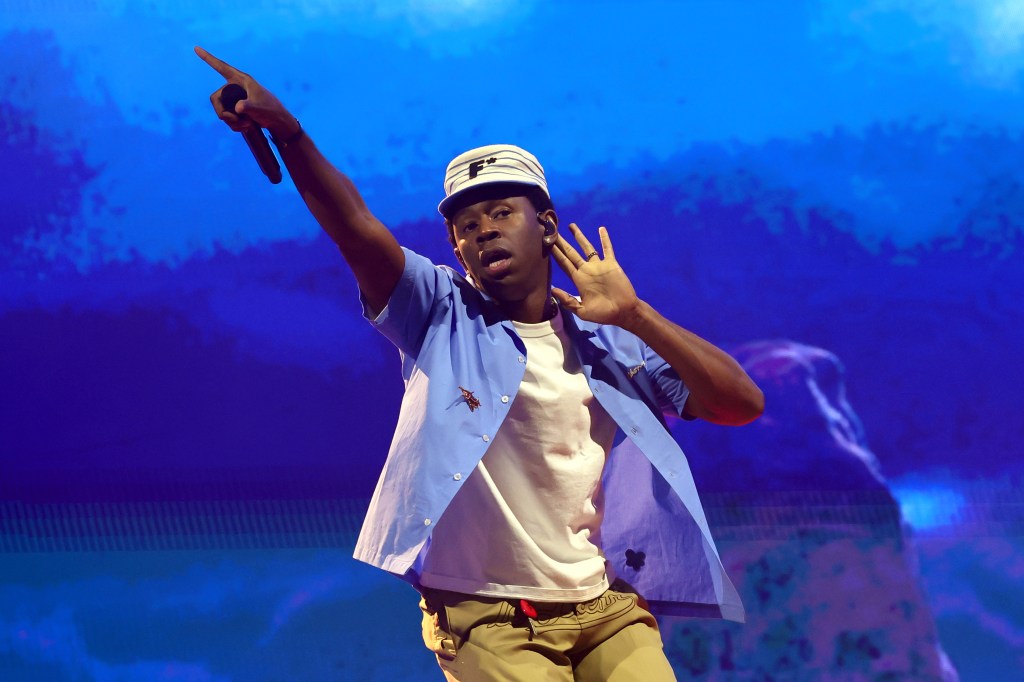 Tyler the Creator performs at the Coachella Stage. He's wearing a blue shirt and white t-shirt, holding his hand to his ear to hear the crowd.