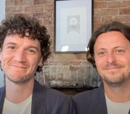 Fiongal and Jamie Greenlaw-Meek both wearing white t-shirts and grey over shirts sat against a brick wall with a moon painting.