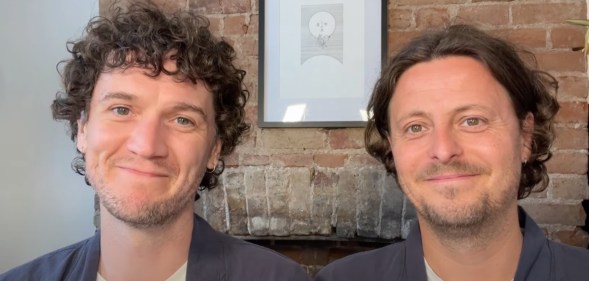 Fiongal and Jamie Greenlaw-Meek both wearing white t-shirts and grey over shirts sat against a brick wall with a moon painting.