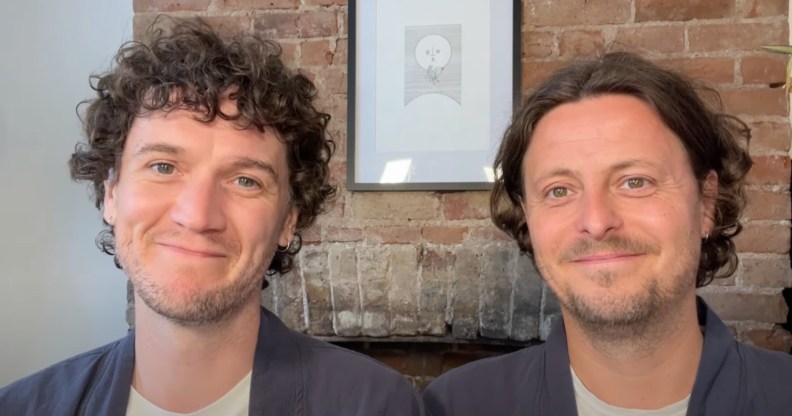 Fiongal and Jamie Greenlaw-Meek both wearing white t-shirts and grey over shirts sat against a brick wall with a moon painting.