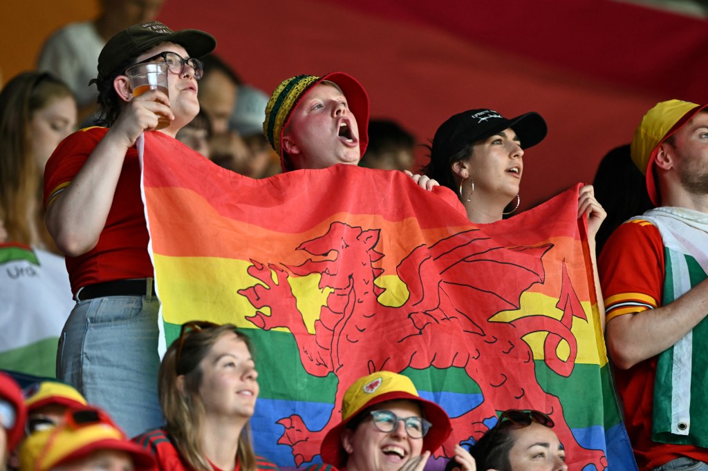Welsh supporter cheer before the start of the UEFA Women's Euro 2025 Group D football match between Wales and The Netherlands. The three fans are holding a Wales flag with rainbow Pride flag colours.