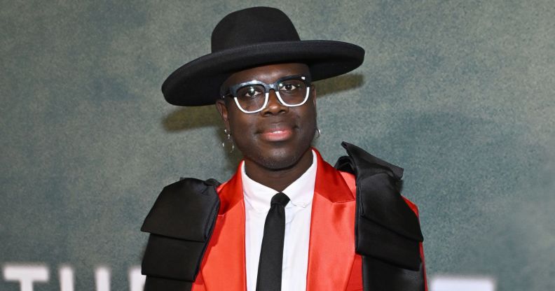 Drag Race star Bob The Drag Queen smiles wearing a black hat, red suit jacket, white shirt and black tie.