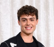 Bradley Riches smiling at the camera. He's wearing a black suit and standing in front of a white curtain