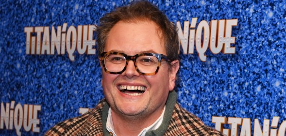 Carr attends the press night performance of "Titanique" at The Criterion Theatre He's wearing a brown stripped coat over a green jumper and white shirt. He's smiling at the camera with turtle-shell brown glasses.