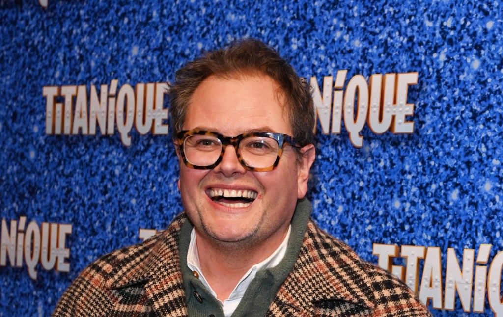 Carr attends the press night performance of "Titanique" at The Criterion Theatre He's wearing a brown stripped coat over a green jumper and white shirt. He's smiling at the camera with turtle-shell brown glasses.