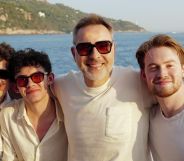 Tobie Donovan, Joe Locke, David Walliams and Kit Connor pose together for a photo with the sea behind them. They are all in white wearing sunglasses, except for Kit Connor, with no sunglasses.