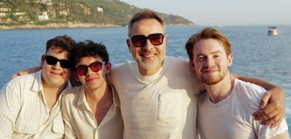 Tobie Donovan, Joe Locke, David Walliams and Kit Connor pose together for a photo with the sea behind them. They are all in white wearing sunglasses, except for Kit Connor, with no sunglasses.