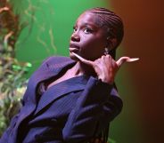 Doechii leans back while wearing a suit and makes a phone gesture with her hand.