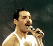 Freddie Mercury performing live on stage. He's singing into the mic wearing a white vest.