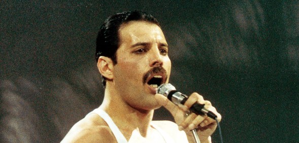 Freddie Mercury performing live on stage. He's singing into the mic wearing a white vest.