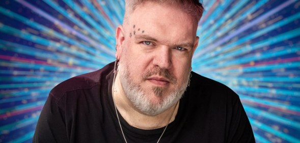 Kristian Nairn wearing a black t-shirt looking into camera with classic Strictly blue sparkles behind him.