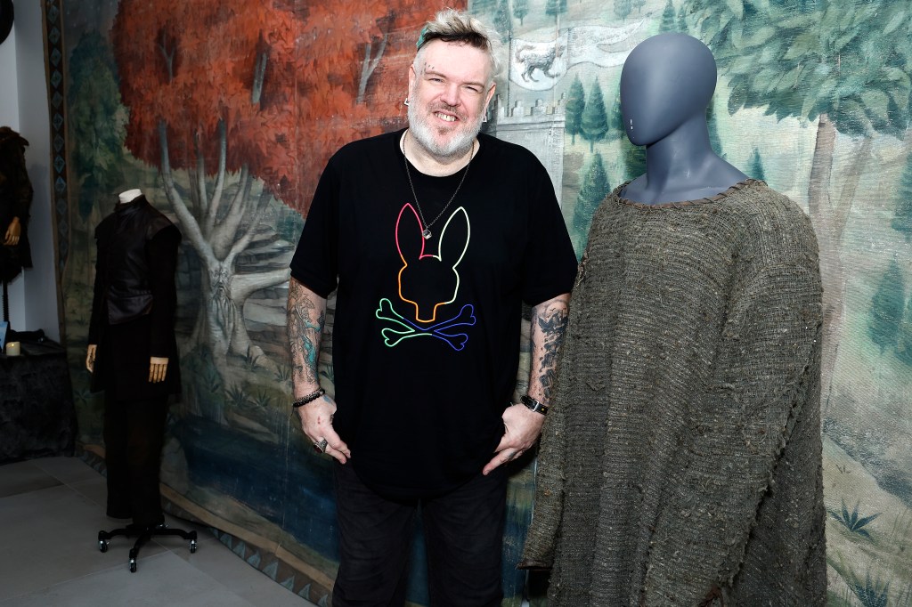 : Kristian Nairn attends the "Game Of Thrones" Mother Of All Auctions at Heritage Auctions. He's wearing a multicoloured t-shirt with a rabbit outline and bones and crosses.