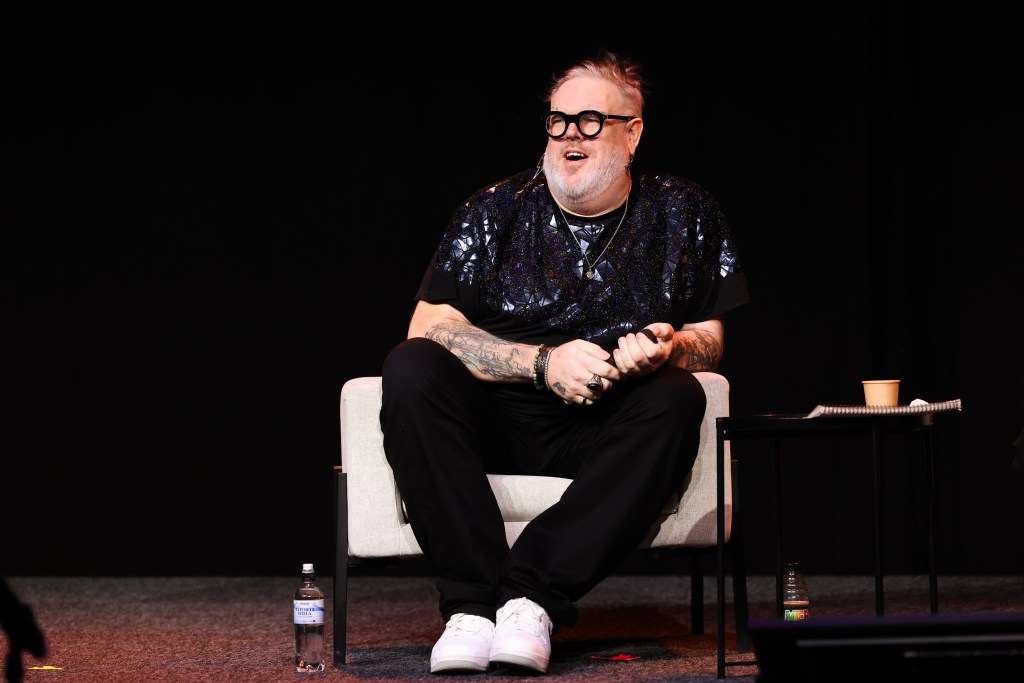 Kristian Nairn sat on a white chair at a panel. He's holding a microphone and wearing a reflective shirt
