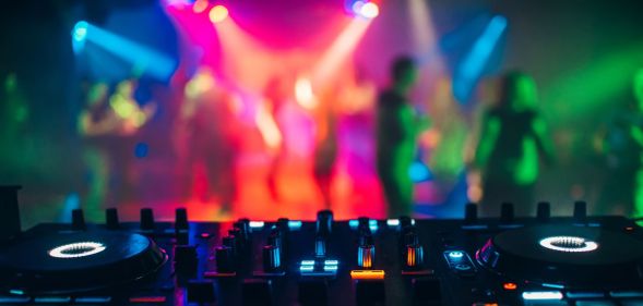 An image of a DJ deck with people dancing under coloured lights in the background.
