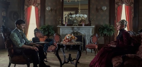 Oscar and Agnes are sat in an ornate living room on chairs opposite eachother, a pot of dying flowers in between them.
