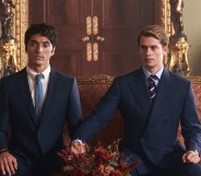 Red, White & Royal Blue still: Alex (Perez) and Prince Henry (Galitzine) sat on a royal red and gold sofa, both wearing suits. Henry has stretched his arm to hold Alex's hand.