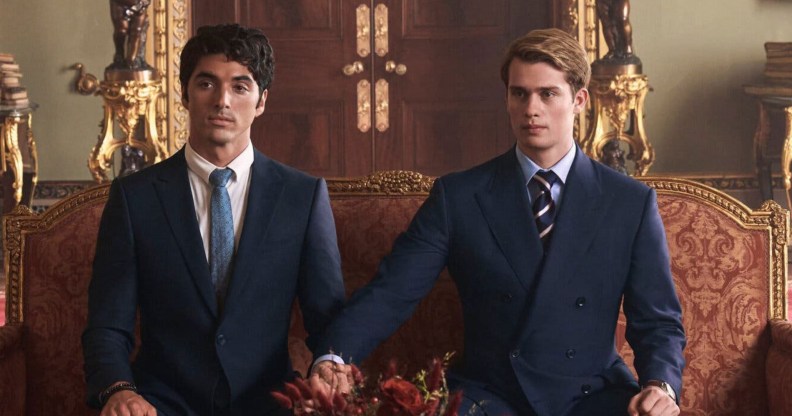 Red, White & Royal Blue still: Alex (Perez) and Prince Henry (Galitzine) sat on a royal red and gold sofa, both wearing suits. Henry has stretched his arm to hold Alex's hand.