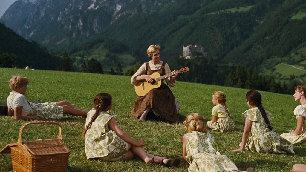 The Sound of Music still: maria with a guitar singing to the children lying on the grassy hill, mountains behind them.