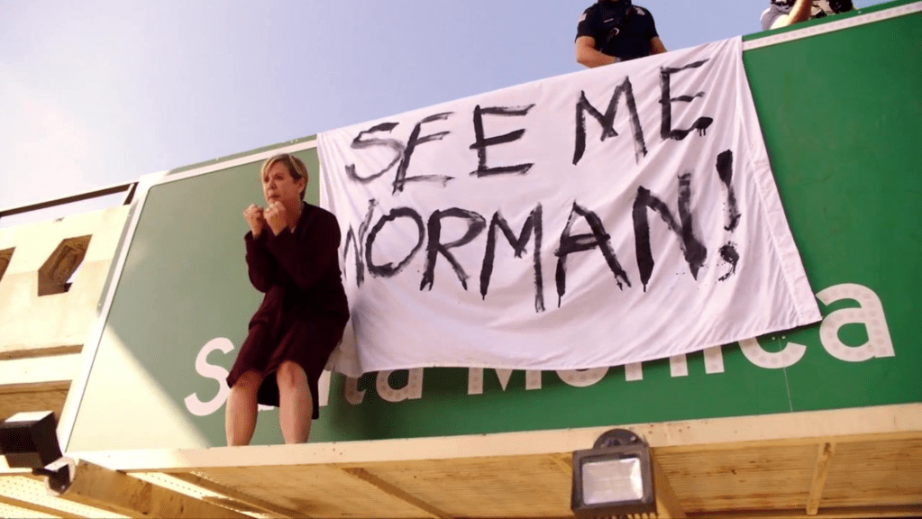 A woman stood on a sign above a freeway with a banner reading 'SEE ME NORMAN'.