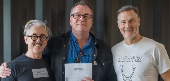 A photo of Russell T Davies (C) smiling and holding a script for Tip Toe, with his arm around actors Alan Cumming (L) and David Morrissey.