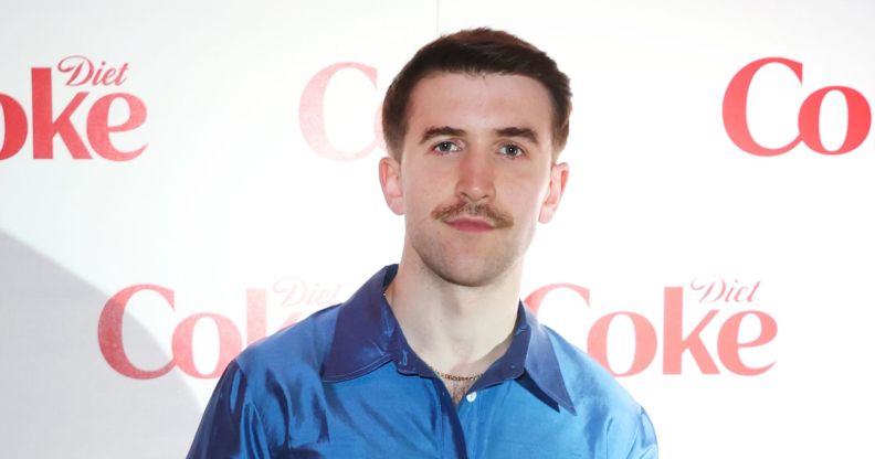 Callum Scott Howells poses in a blue shirt against a white background with Coke written on it.