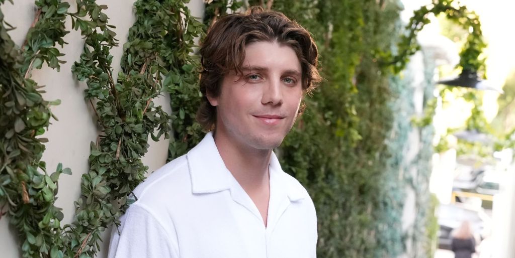 Lukas Gage smiles in a white shirt and poses against a wall with green leaves on it.