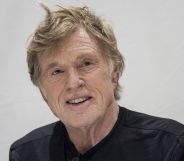 Robert Redford smiles while wearing a black shirt and sitting against a grey background.