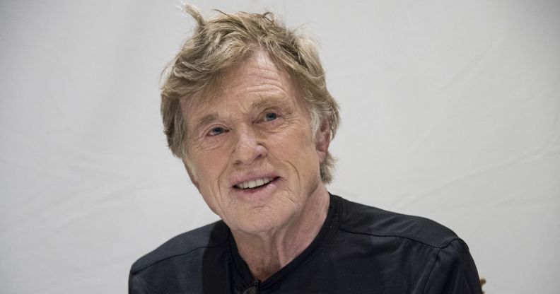 Robert Redford smiles while wearing a black shirt and sitting against a grey background.