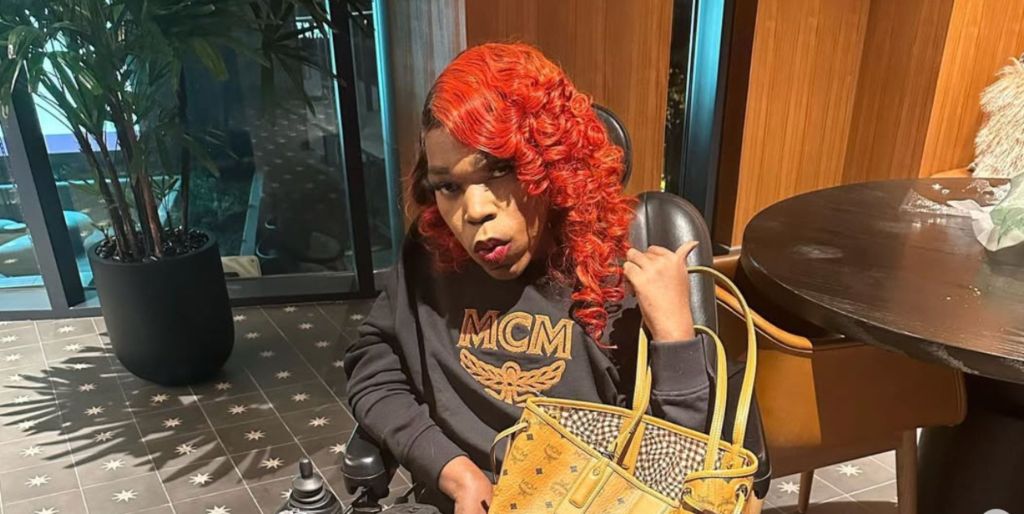 A photo of Rolling Ray with red hair wearing a black jumper with gold embellishments and holding a yellow bag.