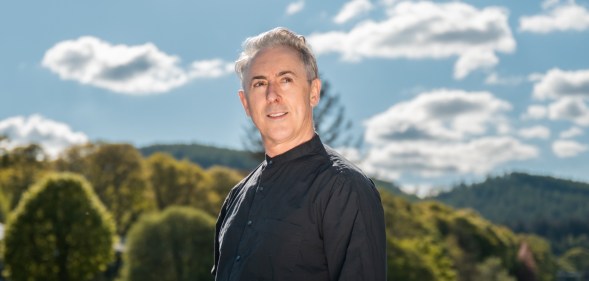 Alan Cumming on Pitlochry Festival Theatre's Out in the Hills. Akab Cumming in a black shirt looking off into the distance, sceneic green view behind him.