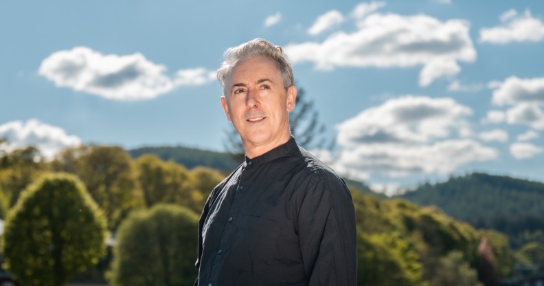 Alan Cumming on Pitlochry Festival Theatre's Out in the Hills. Akab Cumming in a black shirt looking off into the distance, sceneic green view behind him.