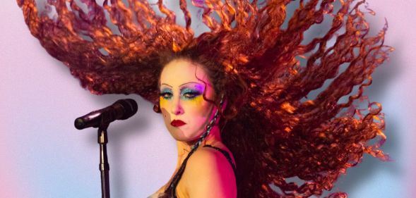Chappell Roan stands by a microphone with her hair in the air. She is against a pink, blue and white background.