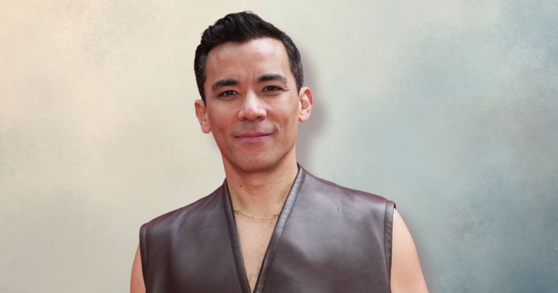 Conrad Ricamora in a grey sleeveless jacket smiling and posing. He is against a blue/grey/cream background