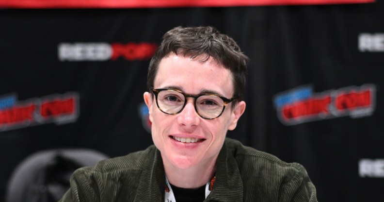 Elliot Page attends New York Comic Con. Eliott wearing a green cordoroy jacket smiling into the camera, wearing round glasses.
