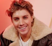 Lukas Gage in a brown leather jacket smiling, sat against an orange background