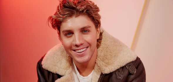 Lukas Gage in a brown leather jacket smiling, sat against an orange background
