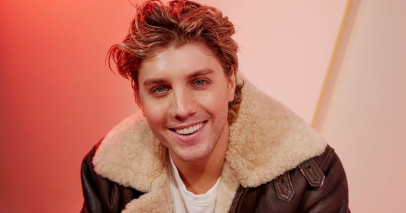 Lukas Gage in a brown leather jacket smiling, sat against an orange background
