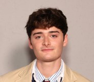 Noah Schnapp in a stripped white and blue shirt with a light beige jacket. Standing against a grey wall