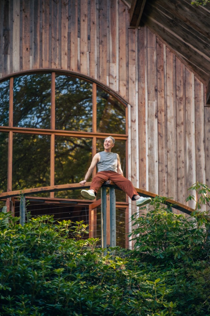 Alan Cumming sat on the end of a balcony fence in front of a wooden house. He's wearing a grey sleeveless best and copper trousers.
