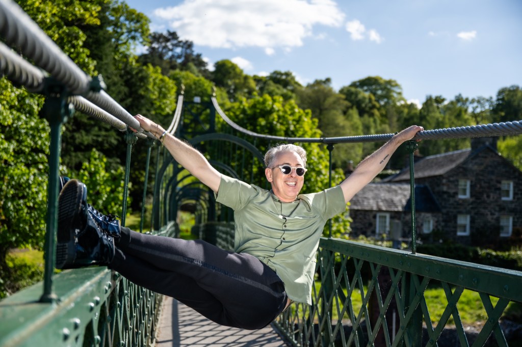 Alan Cumming hanging on a bridge pathway, he's wearing a green shirt and black trousers with green sunglasses.