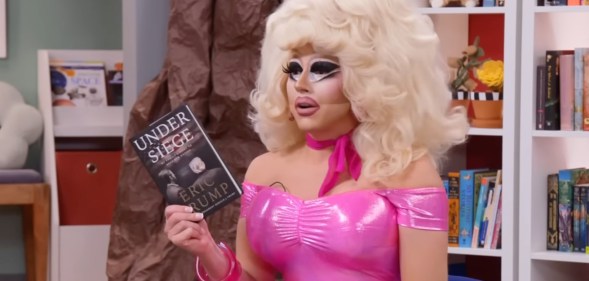 Trixie Mattel in her classic pink dress and big blonde wig holding Eric Trump's book in a school library.